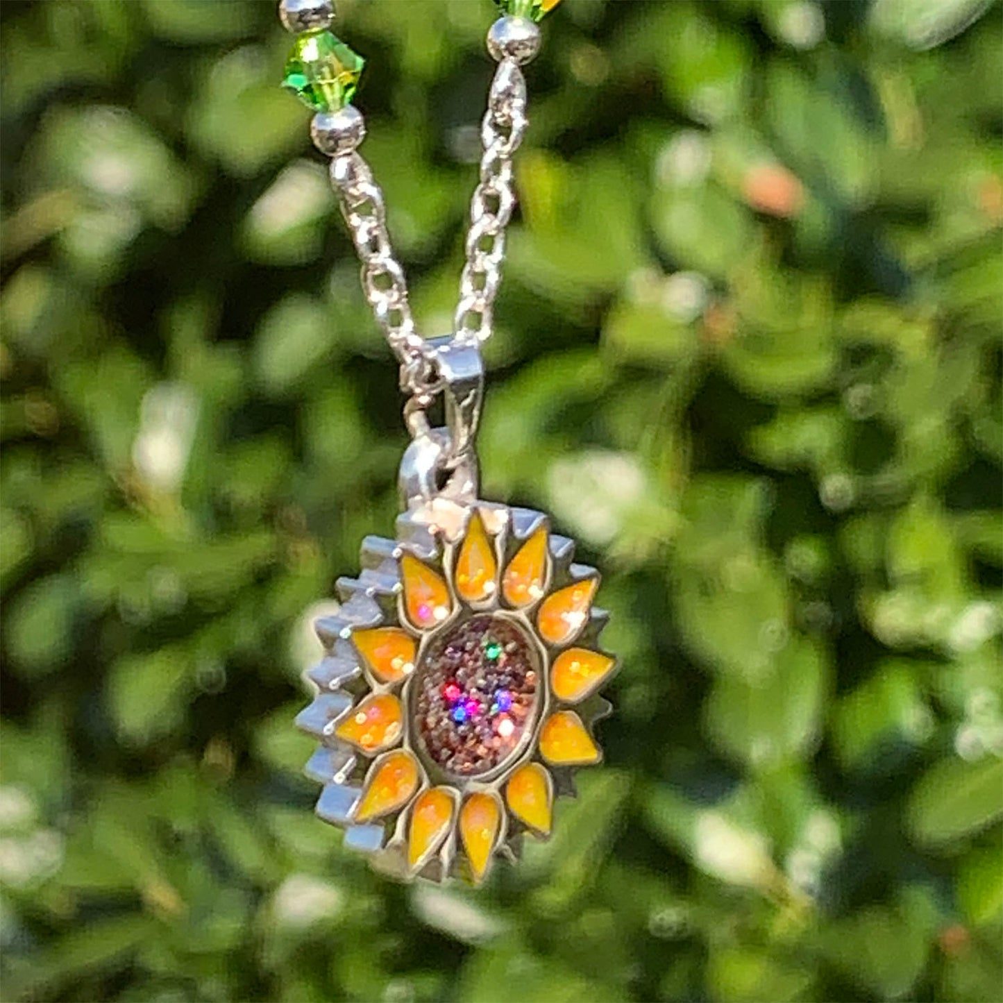 Sunflower pendant with sparkling yellow petals and a round sparkling warm orange centre on a silver long chain with green beads on a green leaf background