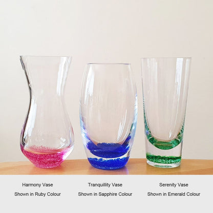 A photograph of three crystal glass memorial vases for ashes in different colours, the Harmony Vase shown in ruby colour, the Tranquillity Vase in sapphire colour, and the Serenity Vase in emerald colour. Each vase has a unique base of colour created by the hand blown process coloured base.