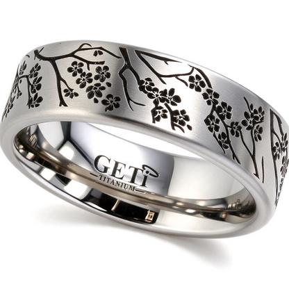 Satin titanium flat-profile silver-coloured ashes ring featuring intricately engraved cherry blossoms and branches of a tree circulating the ring.  Photographed on a white background on a tilted right angle.