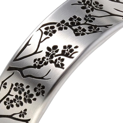 A close-up of a silver-toned metal band featuring an engraved black cherry blossom branch pattern, set against a plain white background.