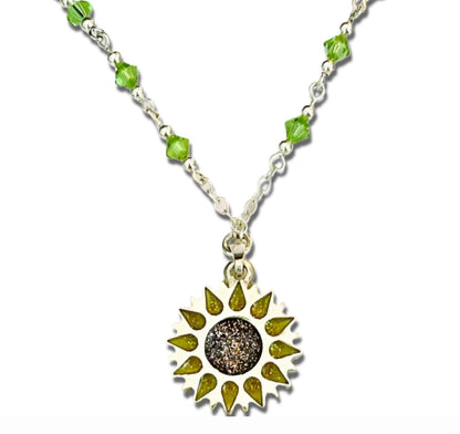 Enlarged close up image of Sunflower pendant with sparkling yellow petals and a round sparkling warm orange centre on a silver long chain with green beads on a white background