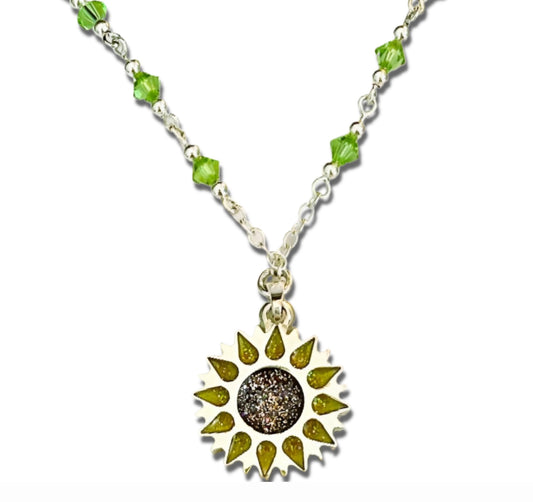 Enlarged close up image of Sunflower pendant with sparkling yellow petals and a round sparkling warm orange centre on a silver long chain with green beads on a white background