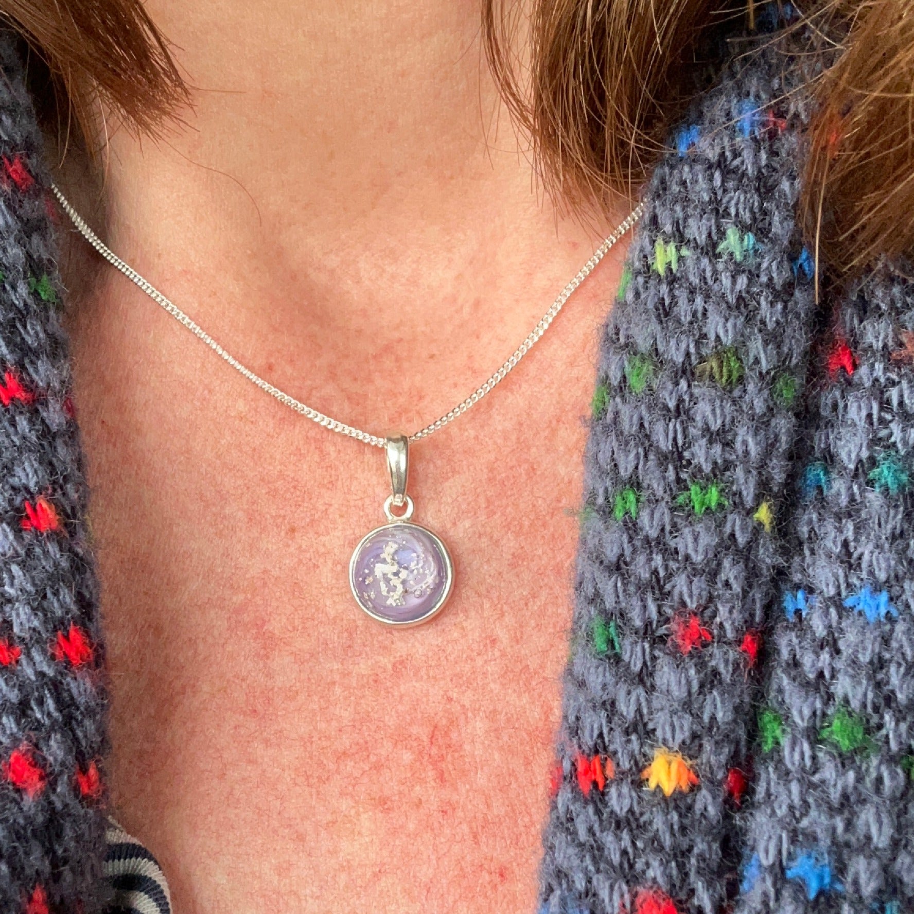 Close-up of a person wearing a silver memorial glass necklace with a round purple charm against a colorful knitted garment.