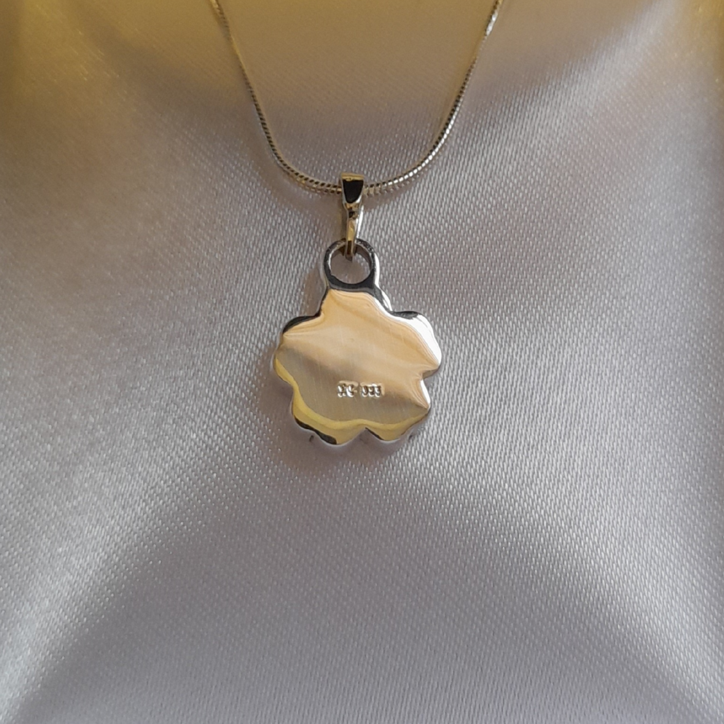 The reverse side of a silver forget me not flower-shaped charm for ashes on a delicate silver chain, displayed on a textured white satin fabric background.