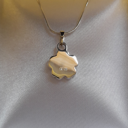The reverse side of a silver forget me not flower-shaped charm for ashes on a delicate silver chain, displayed on a textured white satin fabric background.