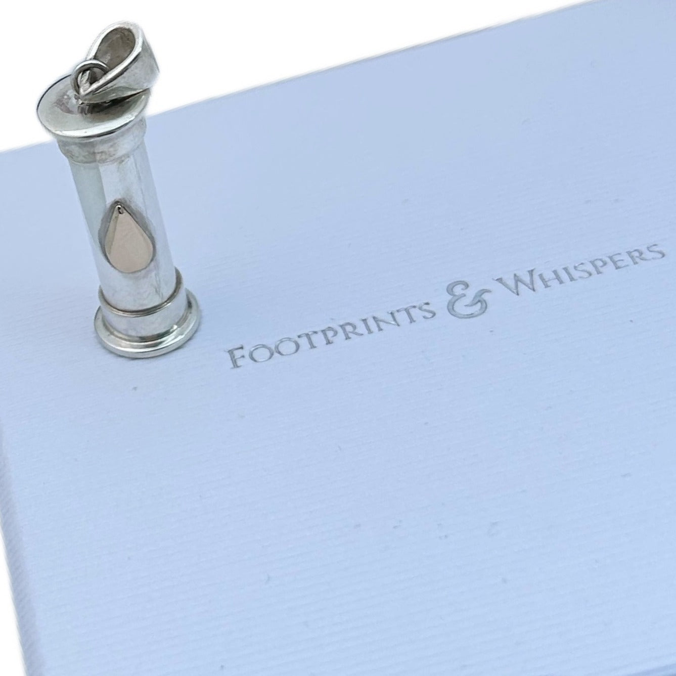 Sterling silver cylindrical self-fill locket with gold teardrop design presented on white footprints and whispers jewellery box